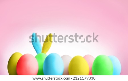 An Easter bunny with blue and yellow ears hides behind painted eggs on a pink background with copy space.
