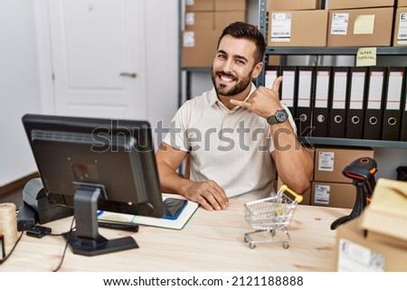 Handsome hispanic man working at small business commerce smiling doing phone gesture with hand and fingers like talking on the telephone. communicating concepts. 