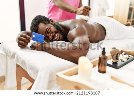 Young african american man reciving herbal pouches thai massage and holding credit card at beauty center.