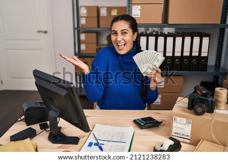 Young hispanic woman working at small business ecommerce holding dollars celebrating achievement with happy smile and winner expression with raised hand 