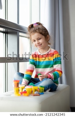 A little girl sits near the window and sculpts from plasticine. She is dressed in a colored jumper. Childhood. Creativity.
