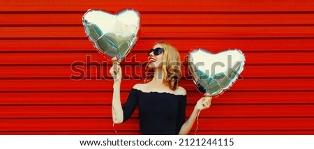 Portrait of beautiful happy smiling woman posing with silver heart shaped balloons on red background