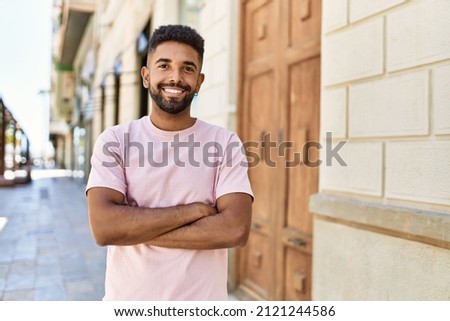 Handsome hispanic man smiling happy with crossed arms outdoors. Young friendly guy standing at the street with cheerful face. Royalty-Free Stock Photo #2121244586