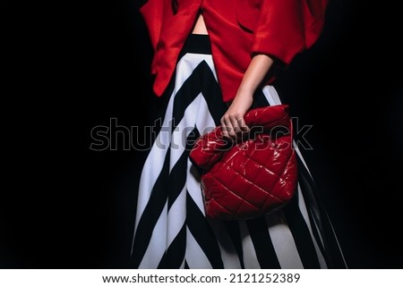 Female figure dressed in a stylish long striped black white skirt and red jacket holding leather creative handbag. Fashion week details