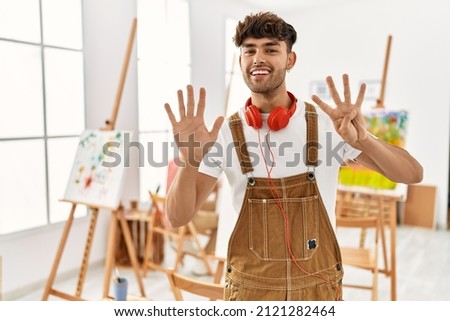 Young hispanic man at art studio showing and pointing up with fingers number nine while smiling confident and happy. 