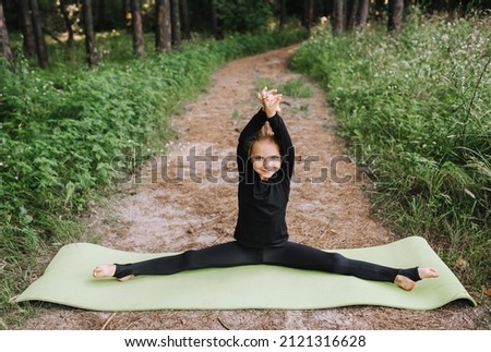 A beautiful, blonde girl, a child, a professional gymnast in a black suit trains, stretches in a twine pose in the woods in nature on a green long rug. Sports photography.