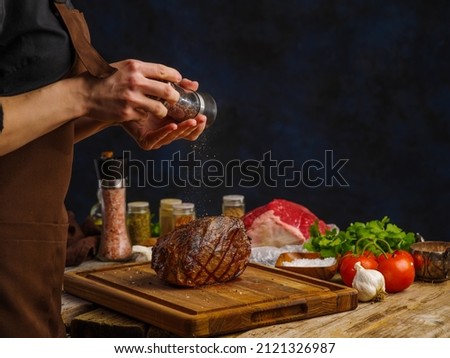 The chef prepares a ham, boiled pork on a wooden cutting board. Sprinkle spices on a piece of cooked meat. Vegetables, herbs, salt, spices. Dark background. The concept is home and restaurant recipes. Royalty-Free Stock Photo #2121326987