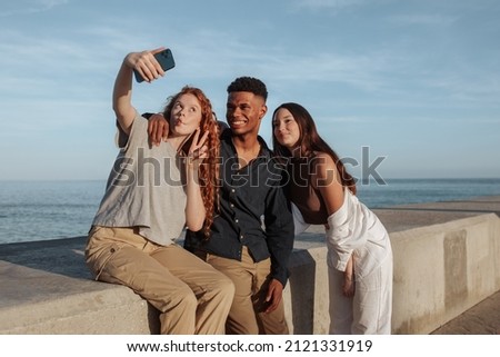 Three young friends taking a group selfie next to the sea. Group of carefree friends posing for the camera phone while hanging out together on the weekend. Friends making memories together.