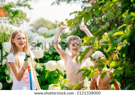 Cute blond girl is pouring a water from a hose at her brothers at backyard cozy garden at country house at summer day. Summer outdoors activity for kids.