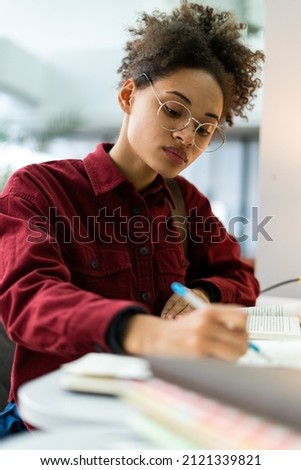 Vertical shot of the focused multiracial school girl studying with books while preparing for test exam or doing homework. Teenage student learning assignment making notes. Teen education concept 