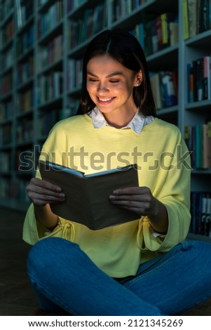 Vertical shot of the young woman reading book at the library and smiling toothy. Brunette lady studying at the high school. Stock photo 