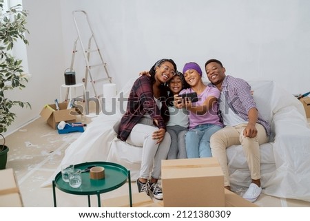 Beautiful girl sits on couch with family in renovated apartment, takes selfie with parents and sister using smartphone, everyone smiles, embraces, poses for photo