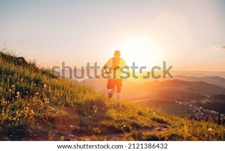 Active mountain trail runner dressed bright t-shirt with backpack running endurance marathon race by picturesque hills at sunset time back view photo. Sporty active people backlight concept image.