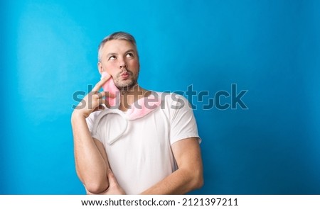 A gay man with makeup in a white T-shirt on a blue background holds rabbit ears in his hands looking at the camera. The concept of LGBT