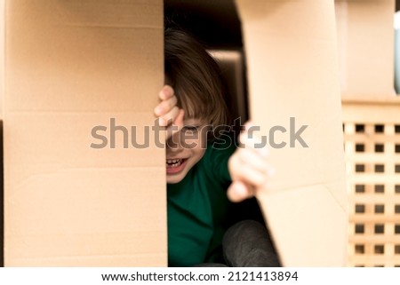 Boy hiding in inside a huge cardboard box. He is playing and peeking through a hole in box. Kid is happy about moving into a new home. Royalty-Free Stock Photo #2121413894