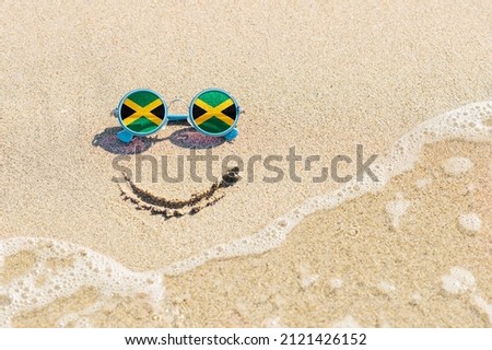 A painted smile on the sand and sunglasses with the flag of Jamaica. The concept of a positive and successful holiday in the resort of Jamaica. Royalty-Free Stock Photo #2121426152