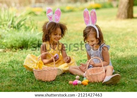 Two children wearing Bunny ears find and pick up multicolored egg on Easter egg hunt in garden Royalty-Free Stock Photo #2121427463
