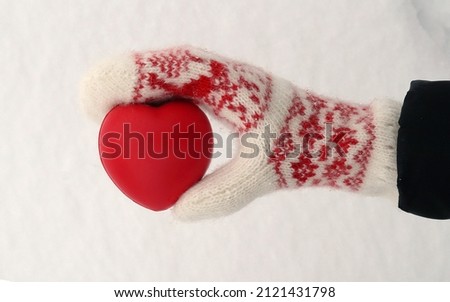 Women's hand in white and red mittens holding a red heart on the snow, copy space