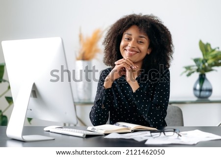 Portrait of young adult successful lovely african american business woman, secretary, sales manager, business consultant, in formal shirt, sitting at workplace with laptop, looking at camera, smiling Royalty-Free Stock Photo #2121462050