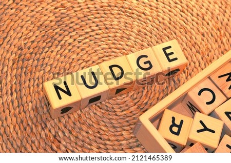 NUDGE word text from wooden cube block letters on braided rattan mats background. Nudge is a verb meaning to push slightly or gently, specifically with an elbow when doing so literally. Royalty-Free Stock Photo #2121465389