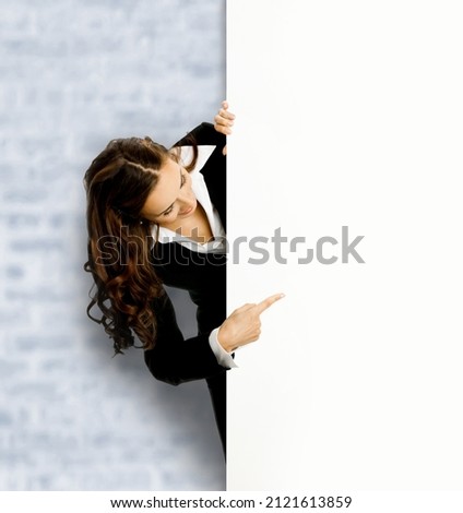 Happy smiling business woman, standing behind, peeping from blank banner or mock up signboard, showing pointing copy space, on white bricks loft wall background