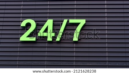 Open seven days a week, 24 hours a day. 24-7 green sign working around the clock hanging on dark wall. Store working 24 hours, sign on facade of building.  
