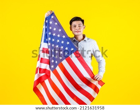 Portrait studio shot of millennial Asian young handsome male US citizen businessman model in casual outfit standing holding big United States of America USA national flag sheet on yellow background.