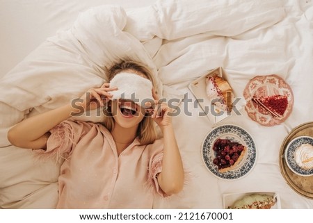 Happy young european girl lies on bed, covers her eyes with sleep mask with open mouth. Blonde in pajamas is enjoying weekend, eating sweets. No healthy food concept Royalty-Free Stock Photo #2121670001
