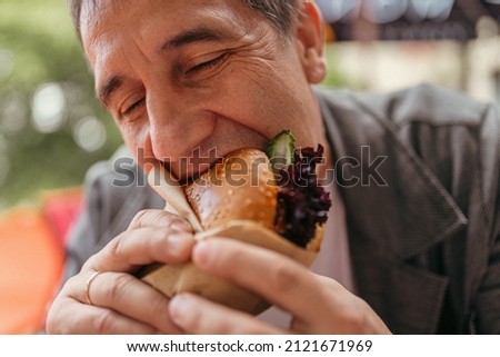 Middle-aged man with appetite eats burger, closing eyes in pleasure. Sitting in fast food restaurant, man has snack Royalty-Free Stock Photo #2121671969