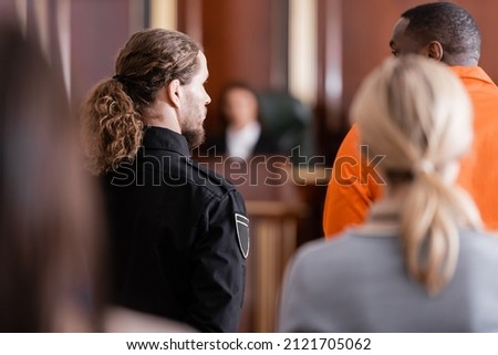 back view of accused african american man near guard and blurred jurors in courtroom Royalty-Free Stock Photo #2121705062
