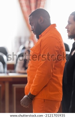 side view of handcuffed african american man in jail uniform near bailiff and blurred jurors in court Royalty-Free Stock Photo #2121705935