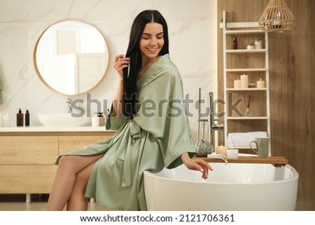 Beautiful young woman sitting on edge of tub in bathroom Royalty-Free Stock Photo #2121706361