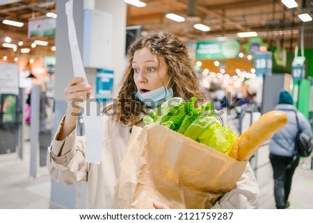 Young woman in a medical mask looks shocked at a paper check in a grocery supermarket holding a paper bag with groceries, price increase and inflation Royalty-Free Stock Photo #2121759287
