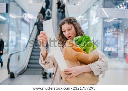 A woman in shock looks into a paper check from a supermarket in a shopping center against the background of an escalator and holds a package with fresh products, price increase Royalty-Free Stock Photo #2121759293
