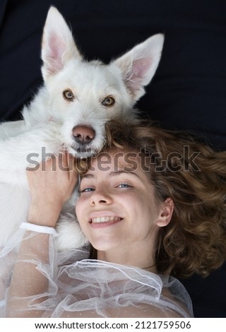 portrait of a cute girl - a teenager of 17-18 years old next to her white dog on a dark background. the similarity and friendship between the female owner and pet. world dog day Royalty-Free Stock Photo #2121759506