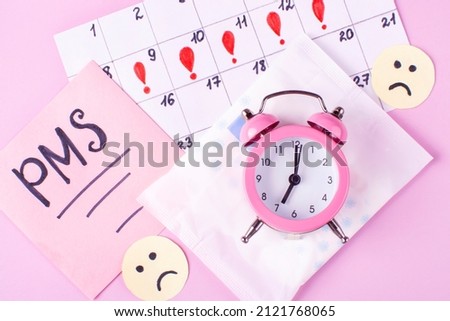 Calendar of women's menstruation, sad emoticons, Menstrual pads (sanitary napkin) in a package, the inscription of PMS, an alarm clock on a pink background. The concept of critical days time. Royalty-Free Stock Photo #2121768065