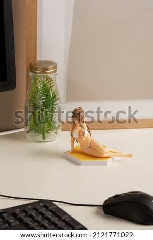Detail shot of a figurine of a long-haired blonde girl in a white bikini. The statuette anime kawaii is located on a computer desk. There is a monitor, a computer mouse and a keyboard on the table.