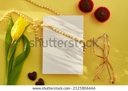 Feminine flat lay with beads with pearls, glasses and chocolate hearts. Trendy shadows and yellow tulip. Beauty postcard concept. Women's Day, happy birthday, anniversary concept. Copy space.