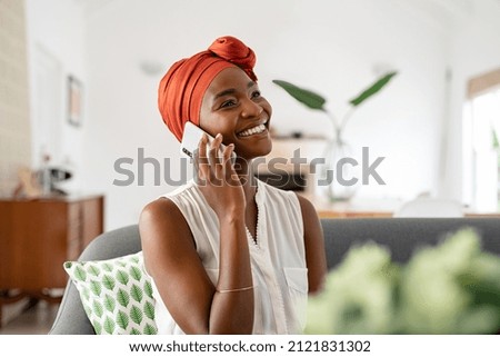 Smiling middle aged woman sitting on couch at home talking over smartphone. Happy black woman with traditional head turban using mobile phone for conversation. Relaxing mid african american lady. Royalty-Free Stock Photo #2121831302