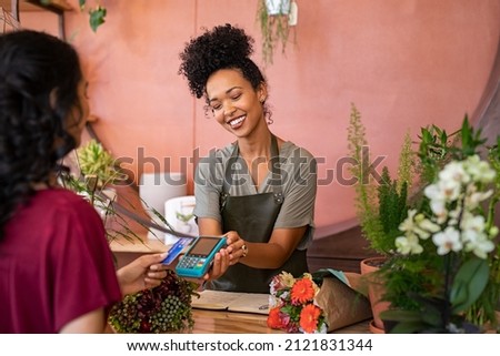 Smiling florist holding card reader machine at counter with customer paying with credit card. Young african american florist shop assistant holding payment machine while buyer purchase a bunch flower. Royalty-Free Stock Photo #2121831344