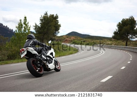 Biker in a black uniform driving a white sports motorcycle on a curvy road in the middle of a green meadow with trees and with mountains in the background on a cloudy day Royalty-Free Stock Photo #2121831740
