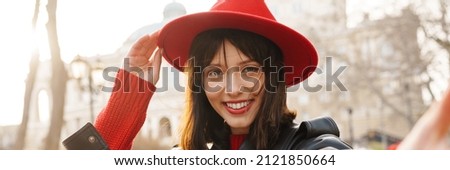 Close up of a smiling young stylish brunette white woman wearing hat and leather jacket with short hair standing on a street taking selfie