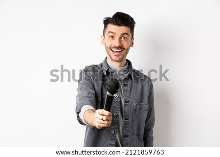 Handsome smiling man stretch out hand with microphone, giving you mic, standing on white background