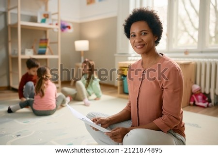 Smiling African American preschool teacher sitting on the floor and looking at camera. There are kids playing in the background.