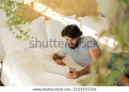 Young man working at home from his bedroom with laptop on his bed. Man lying down. Man lying down. Gray notebook for working. Cozy and comfortable. Home office concept. High quality photo