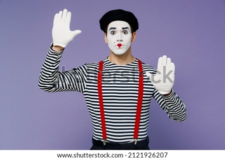 Bright young mime man with white face mask wears striped shirt beret looking camera hands raised as if touch stand near invisible wall isolated on plain pastel light violet background studio portrait