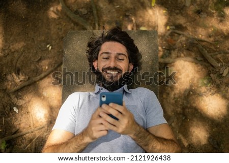 View of young man using a smartphone at day time lying down on a bench at a park. High quality photo. Mobile phone, technology, urban, nature  concept. High quality photo
