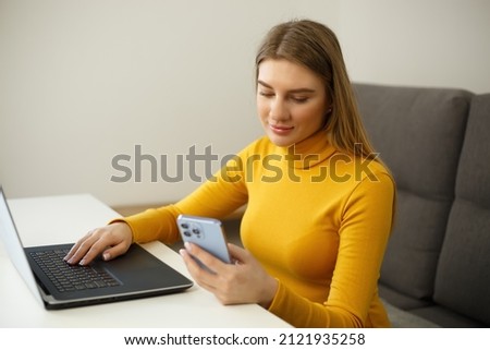 Beautiful young blonde woman using mobile phone and laptop computer for communication and distant work during lockdown. Attractive white female typing message on new smartphone with cheerful smile