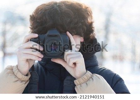 A teenage boy with curly long hair takes a picture with a digital SLR camera. Winter photo shoot outdoors