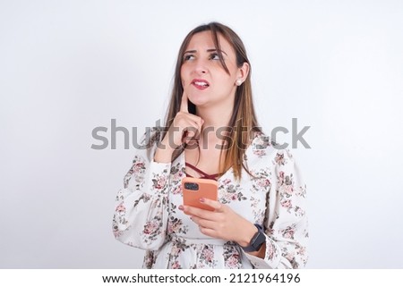 Image of a thinking dreaming Young arab woman wearing floral shirt over white backgtound  using mobile phone and holding hand on face. Taking decisions and social media concept.